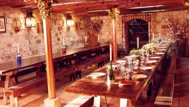 Een lange houten tafel gedekt voor een maaltijd, met banken aan weerszijden, stenen muren en een stenen boog in de achtergrond.