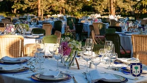 Een buitenterras met meerdere tafels gedekt voor een maaltijd, omringd door rieten stoelen en versierd met bloemen.