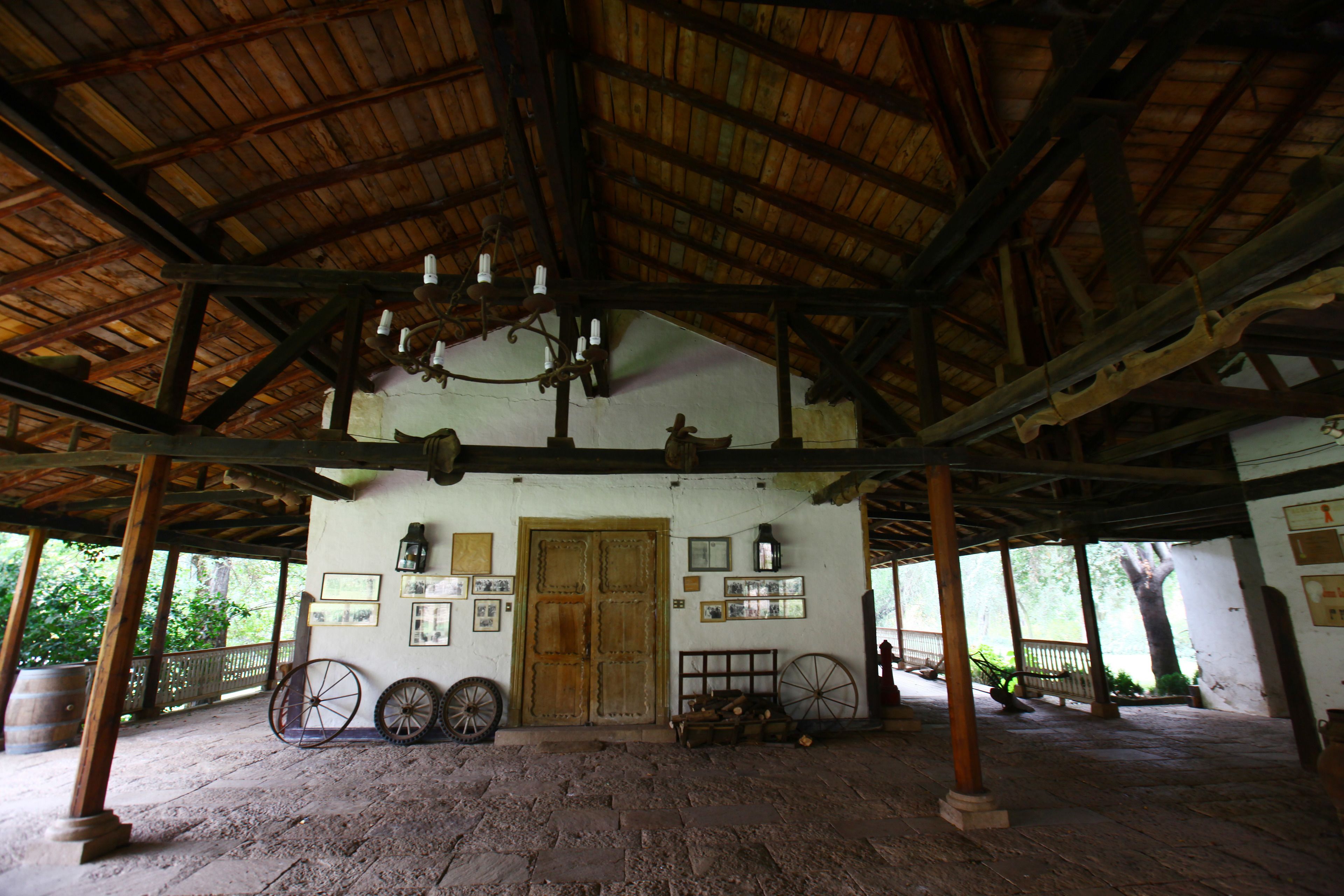 A rustic covered porch with wooden beams, a wooden door, and a chandelier. There are framed pictures on the walls and a wheelbarrow in the center.