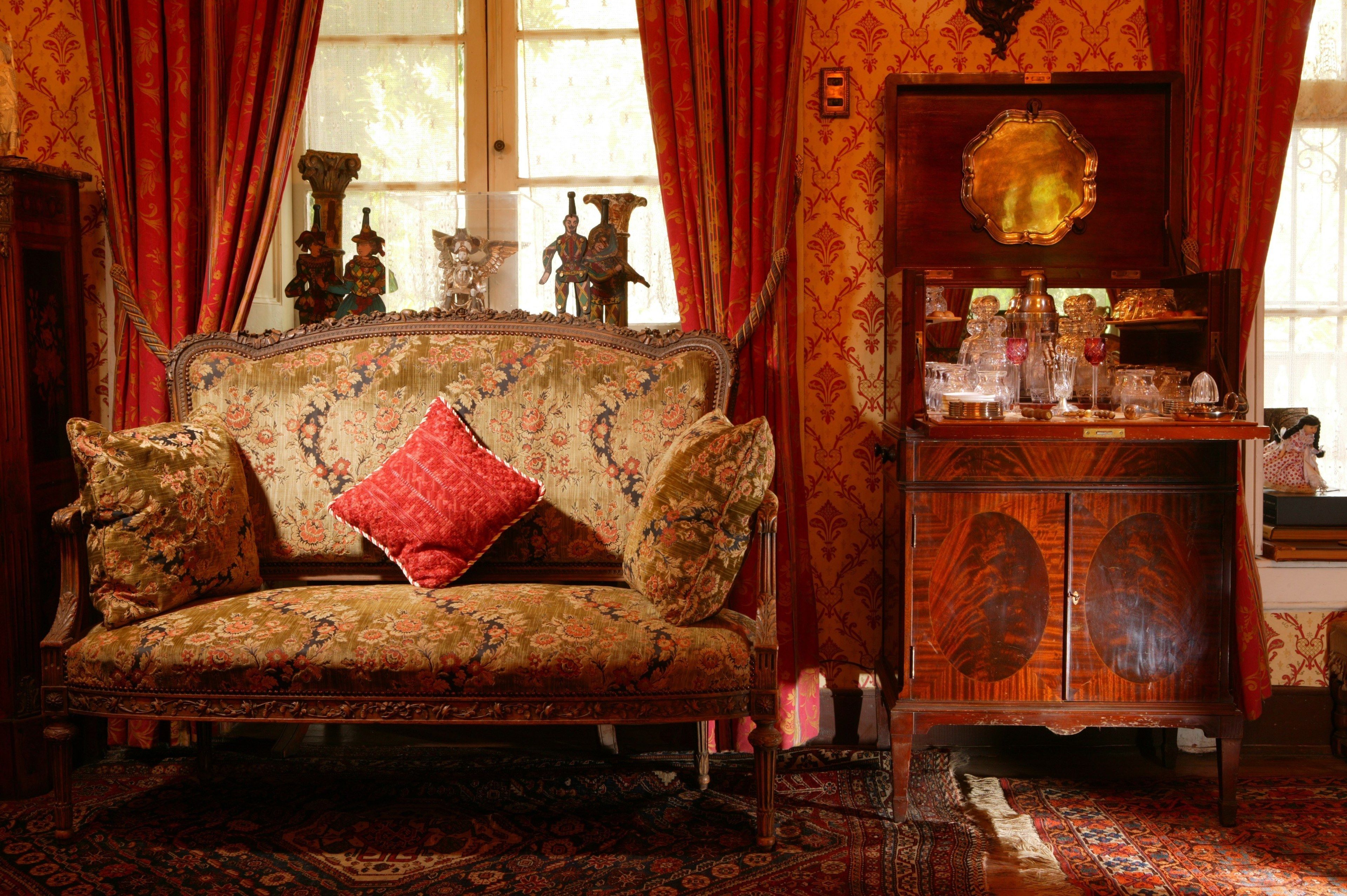 A vintage-style room with a floral-patterned sofa, a wooden cabinet with glassware, and a large mirror.