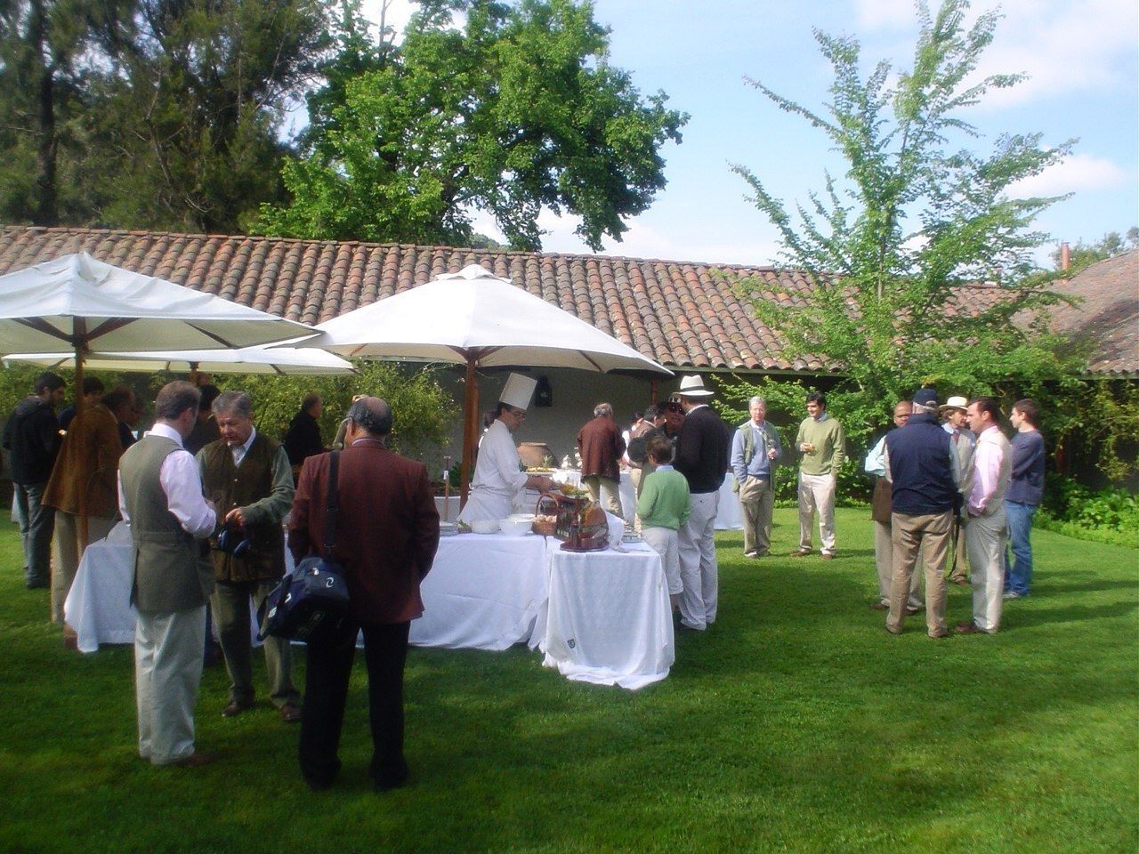 Evento al aire libre con gente reunida alrededor de mesas bajo sombrillas, con un edificio en el fondo.