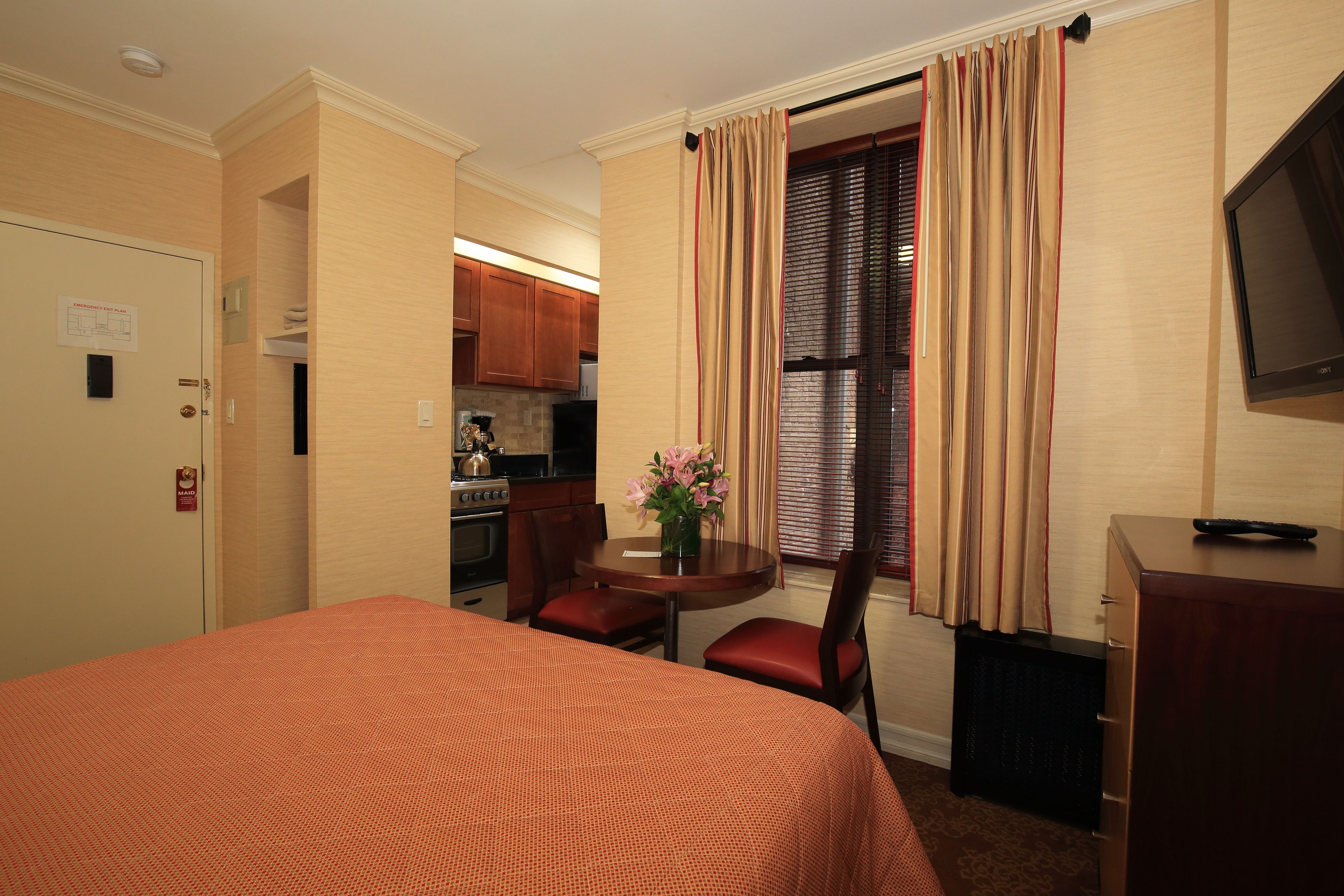 A hotel room with a bed, a desk, a chair, a small table with a vase, and a kitchenette in the background.