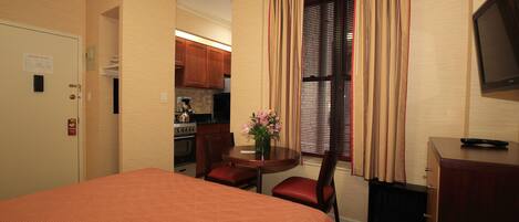 A hotel room with a bed, a desk, a chair, a small table with a vase, and a kitchenette in the background.