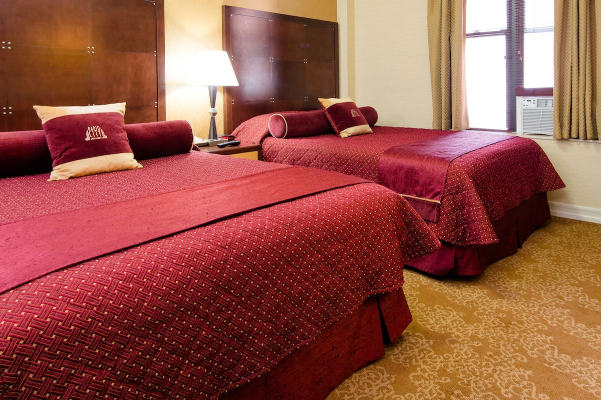 A hotel room with two beds, a nightstand, a lamp, and a window with curtains.