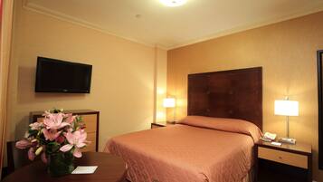 A hotel room with a bed, bedside lamps, a TV, a vase with flowers, and a table with a chair.