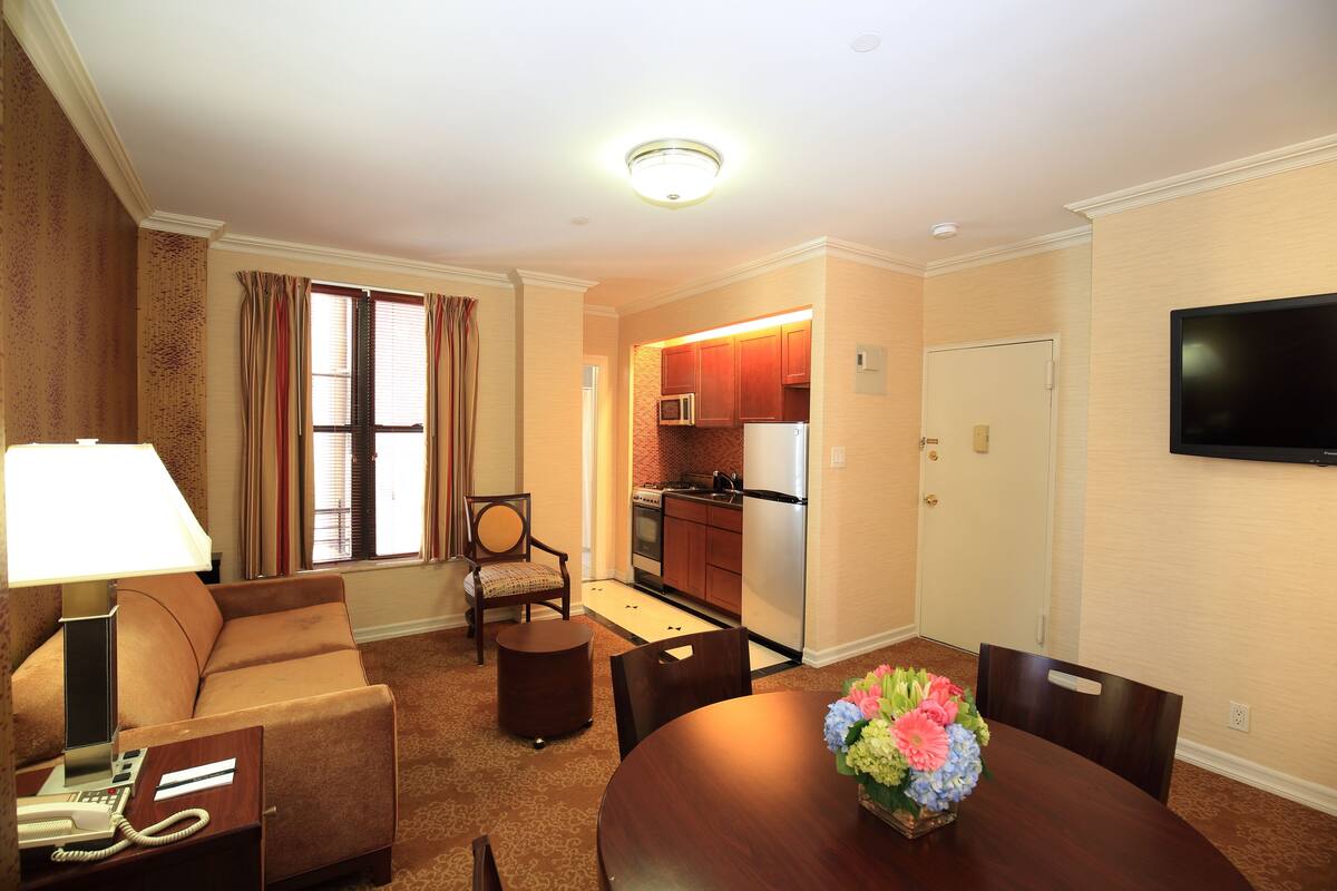 A hotel room with a sofa, a dining table with chairs, a small round table, a lamp, a telephone, a TV mounted on the wall, and a kitchen area with a microwave and refrigerator.