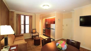 A hotel room with a sofa, a dining table with chairs, a small round table, a lamp, a telephone, a TV mounted on the wall, and a kitchen area with a microwave and refrigerator.