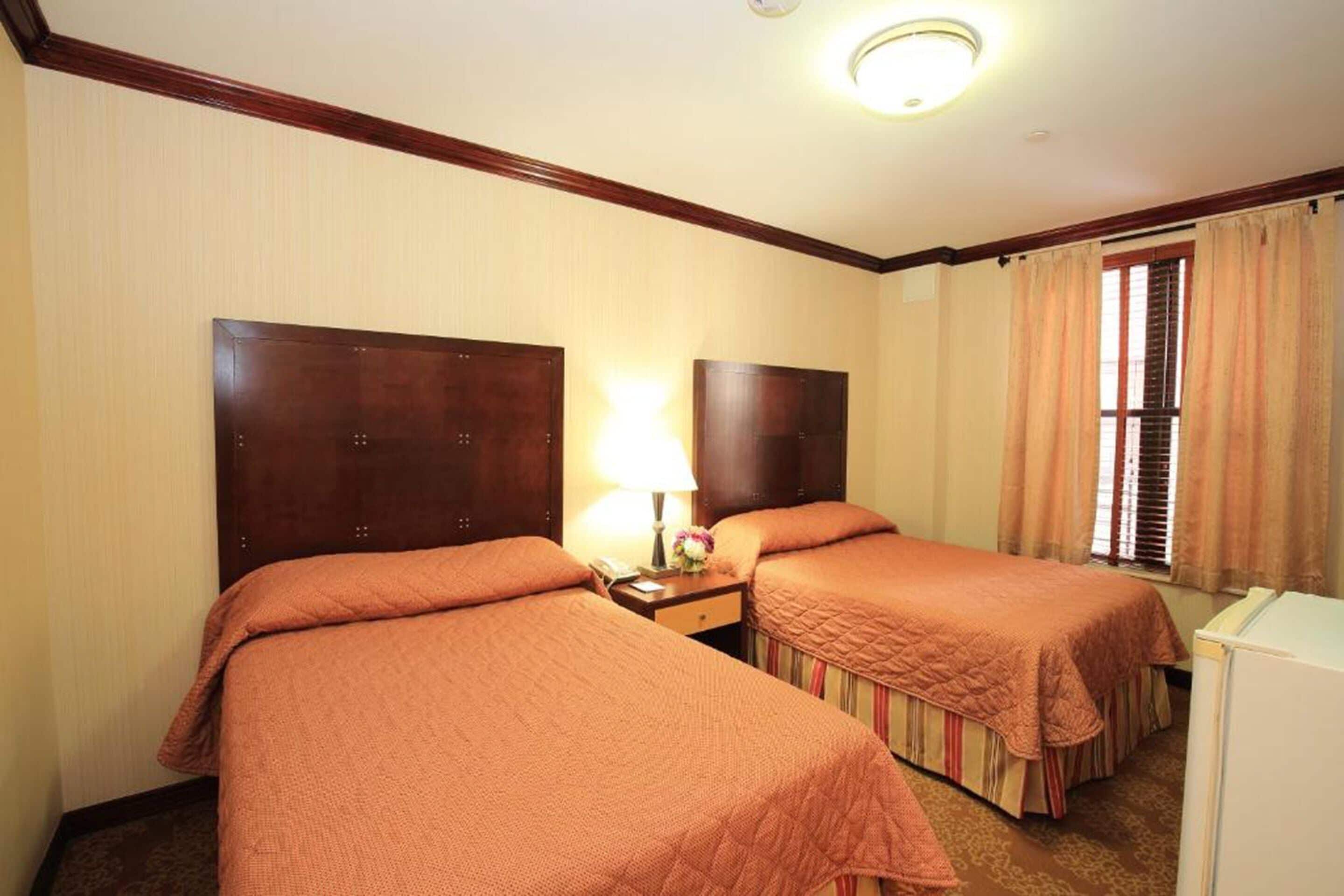 A hotel room with two beds, a nightstand with a lamp, a small table with a vase of flowers, and a window with curtains.