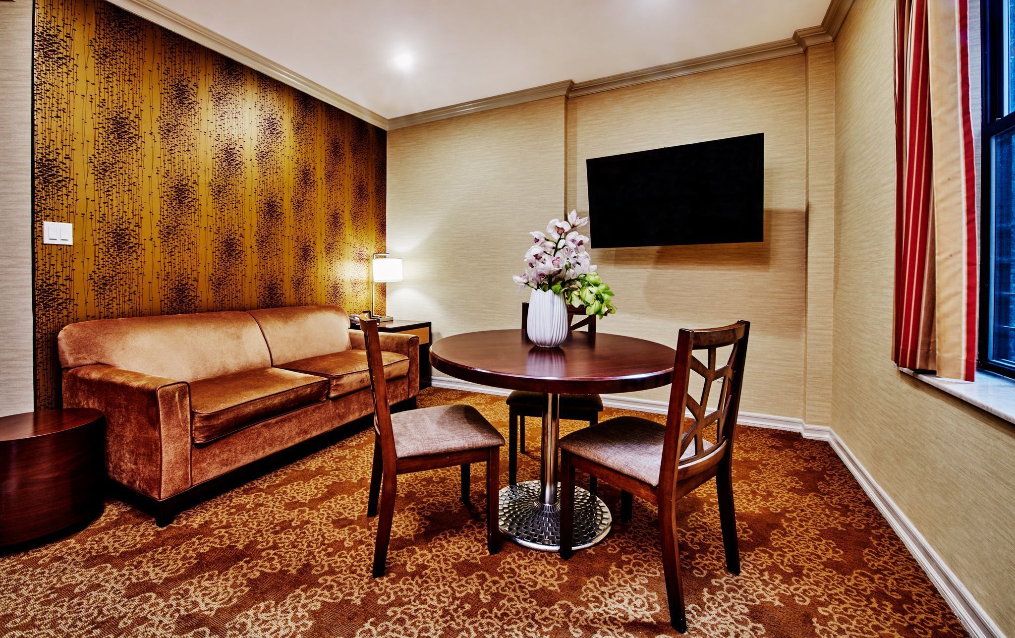 Een hotelkamer met een bruin kamerkleed, een bruine bank, een ronde houten tafel met vier stoelen, een vaas met bloemen, een flatscreen televisie aan de muur en een raam met rode gordijnen.