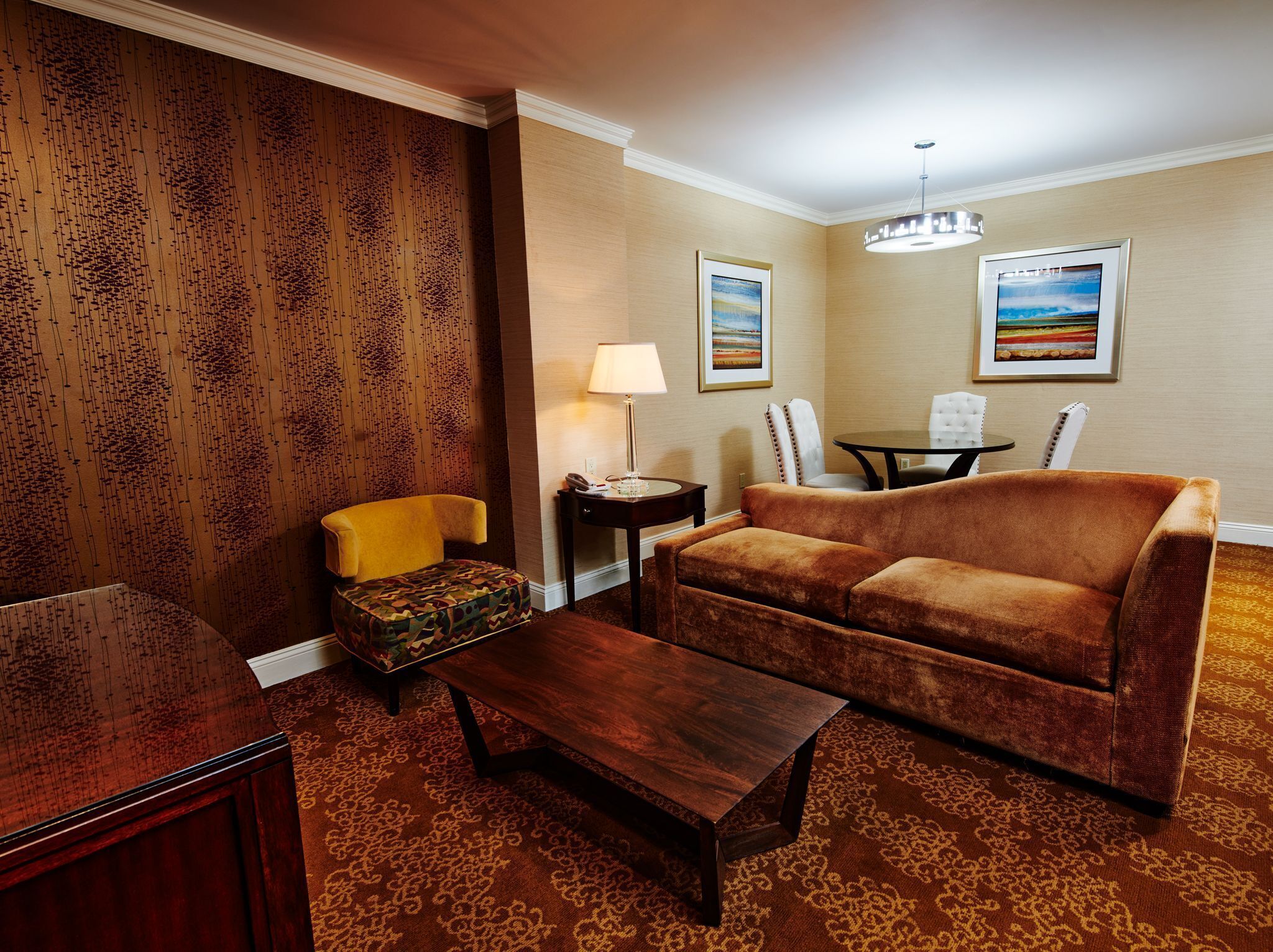 A living room with a brown sofa, a yellow armchair, a patterned ottoman, a wooden coffee table, and a small side table with a lamp. There are two framed pictures on the wall and a ceiling light fixture.