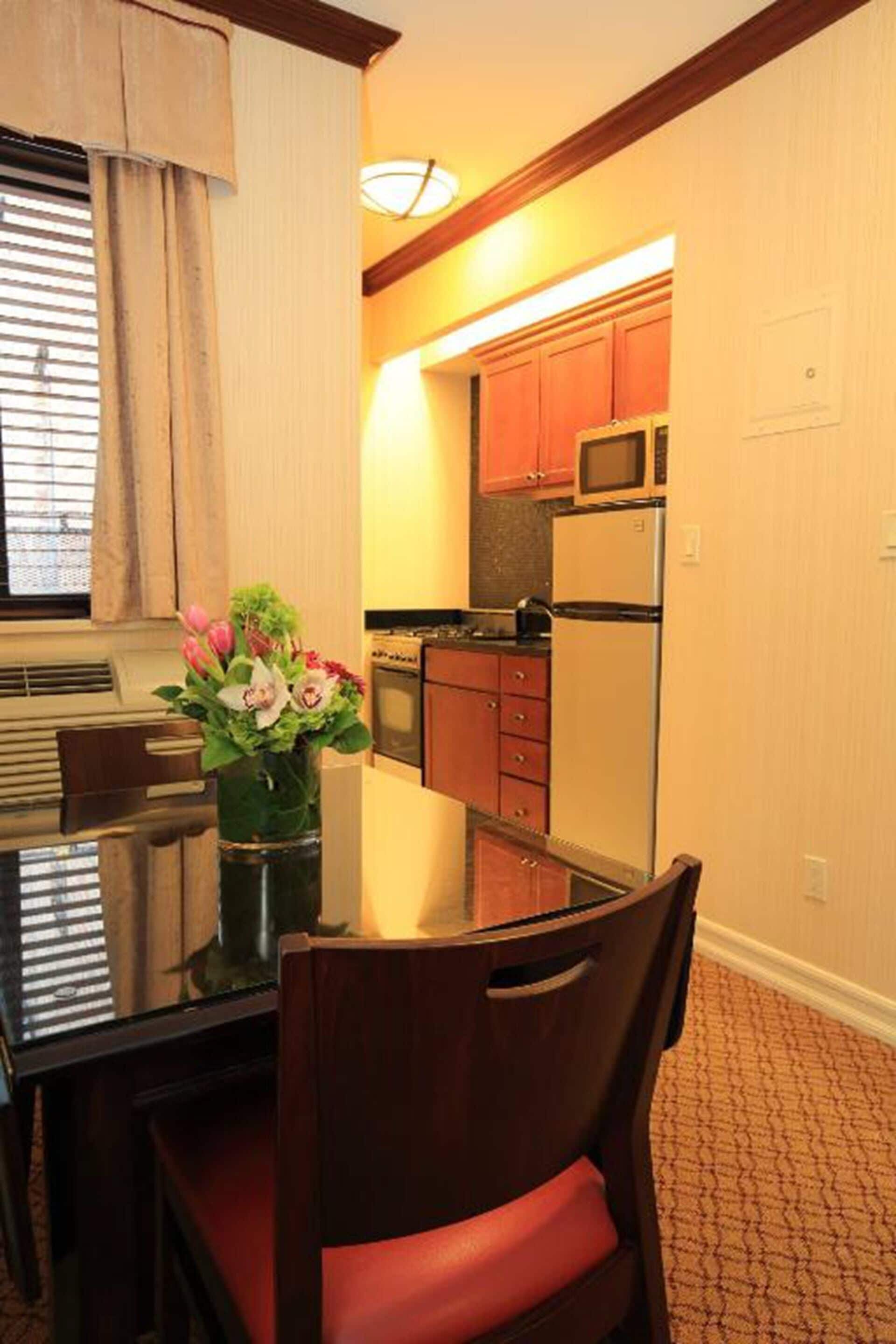 A dining area with a table and chairs, a vase with flowers, and a view into a kitchen with cabinets and appliances.