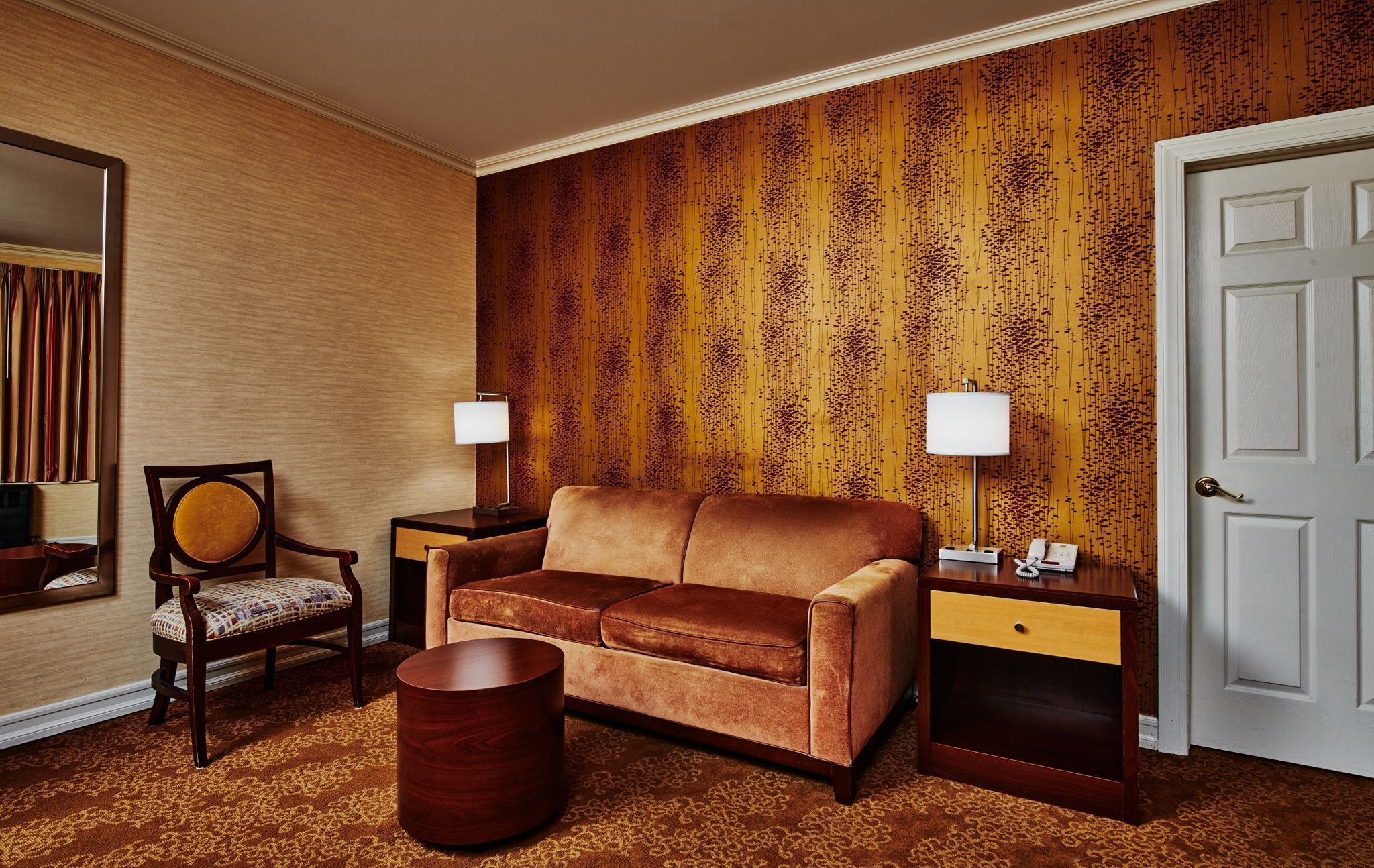 Une chambre d’hôtel avec un canapé marron, une table d’appoint en bois avec une lampe, une chaise en bois, une petite table ronde en bois et un tapis à motifs.