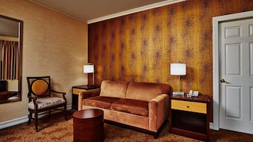 Une chambre d’hôtel avec un canapé marron, une table d’appoint en bois avec une lampe, une chaise en bois, une petite table ronde en bois et un tapis à motifs.