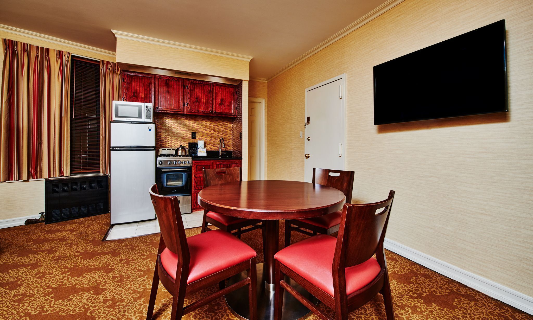 A compact hotel room with a dining table, four chairs, a TV mounted on the wall, a kitchen area with a stove, microwave, and refrigerator, and a patterned carpet.