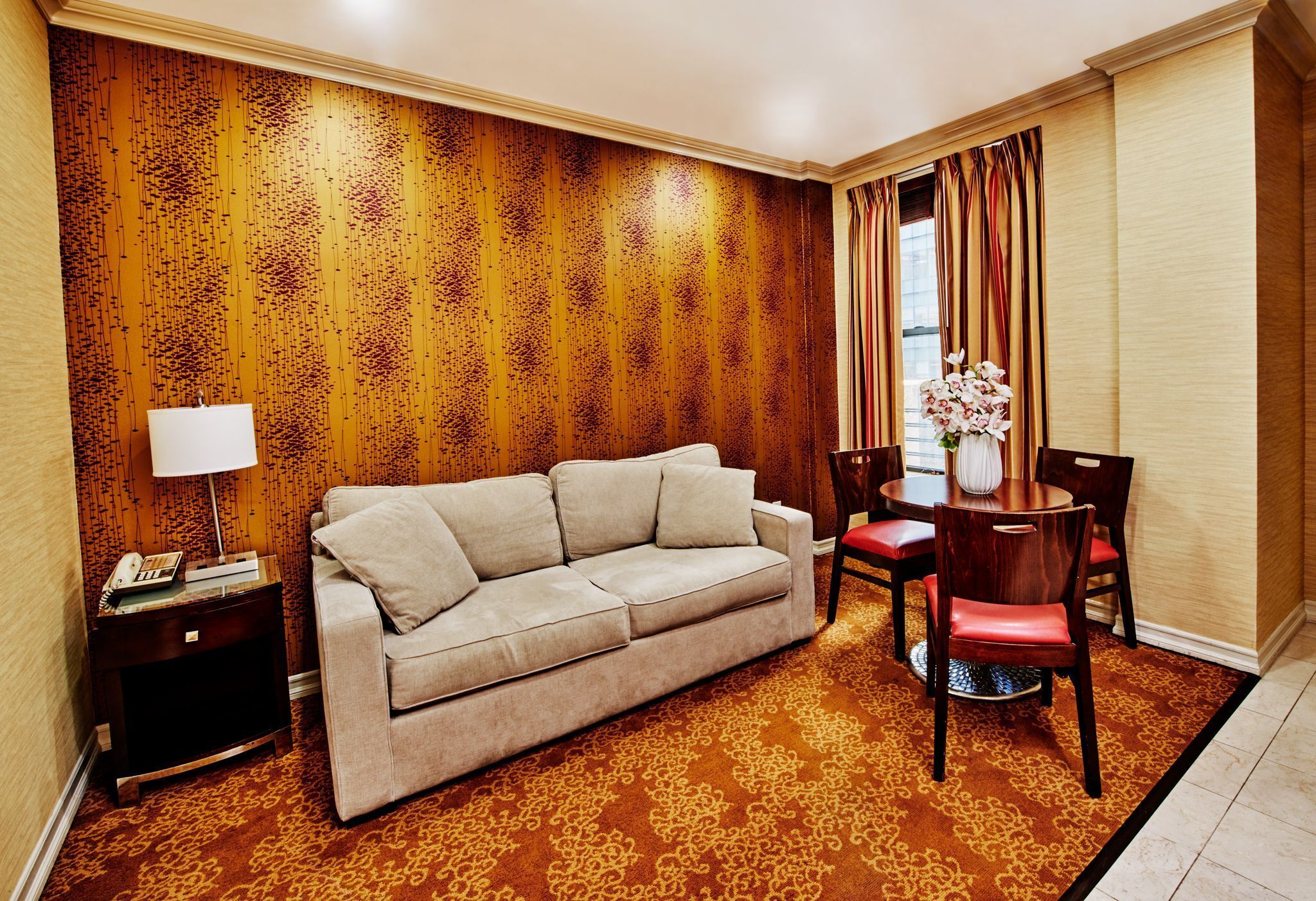 A hotel room with a beige sofa, a wooden accent wall, a patterned carpet, a dining table with chairs, and a vase of flowers.