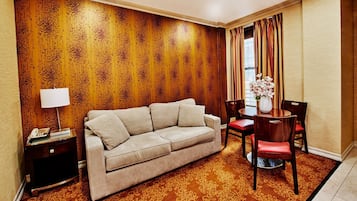 A hotel room with a beige sofa, a wooden accent wall, a patterned carpet, a dining table with chairs, and a vase of flowers.