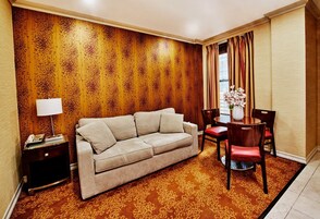Hotelkamer met een beige bank, een houten accentmuur, een kamerbreed tapijt met dessin, een eettafel met stoelen, en een bloemenvaas.