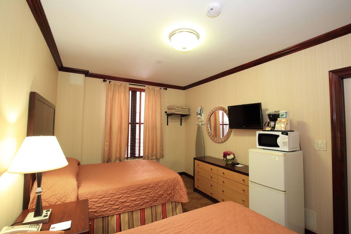 A hotel room with two beds, a desk with a lamp, a dresser, a microwave, and a television on the wall.