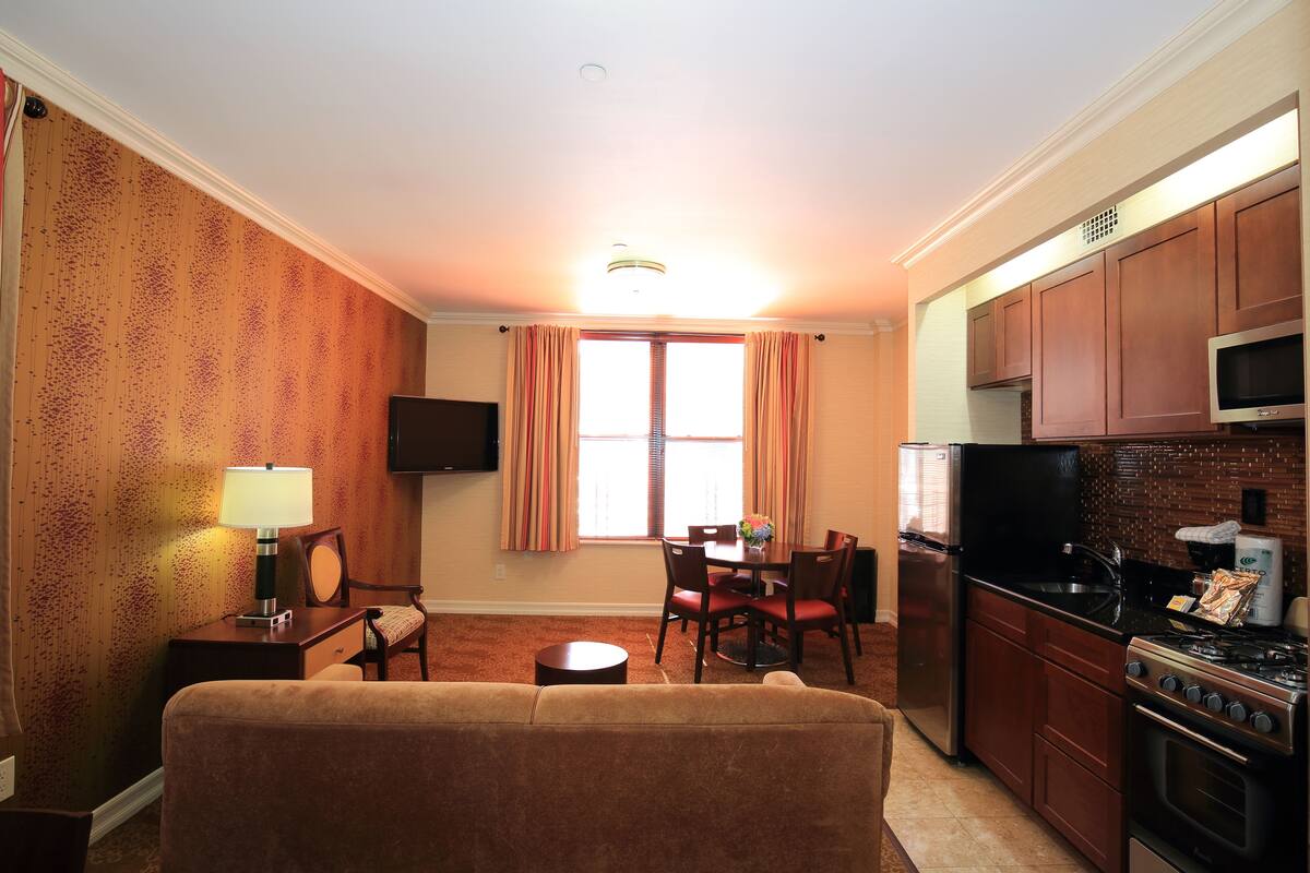 A compact living space with a sofa, dining table, chairs, a TV, a kitchen area with a stove, microwave, and refrigerator.