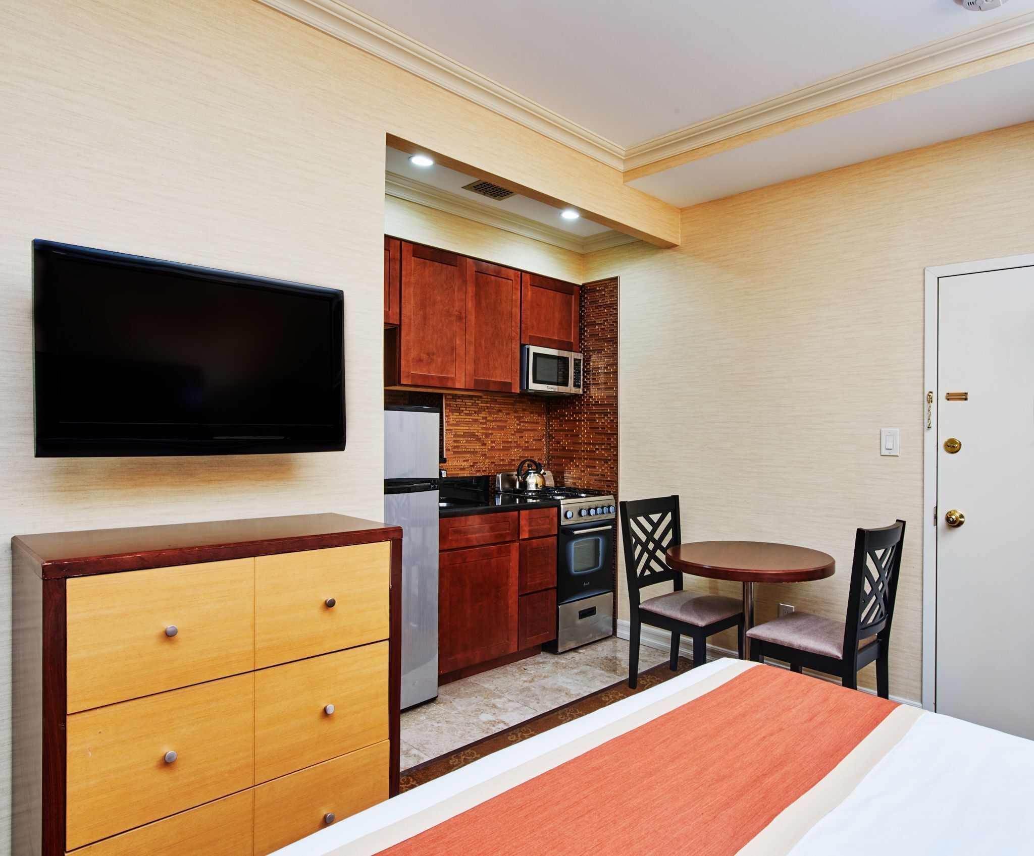 Una habitación de hotel compacta con cama, televisor, kitchenette con microondas, estufa y refrigerador, y un pequeño comedor con mesa y sillas.