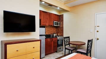 Una habitación de hotel compacta con cama, televisor, kitchenette con microondas, estufa y refrigerador, y un pequeño comedor con mesa y sillas.
