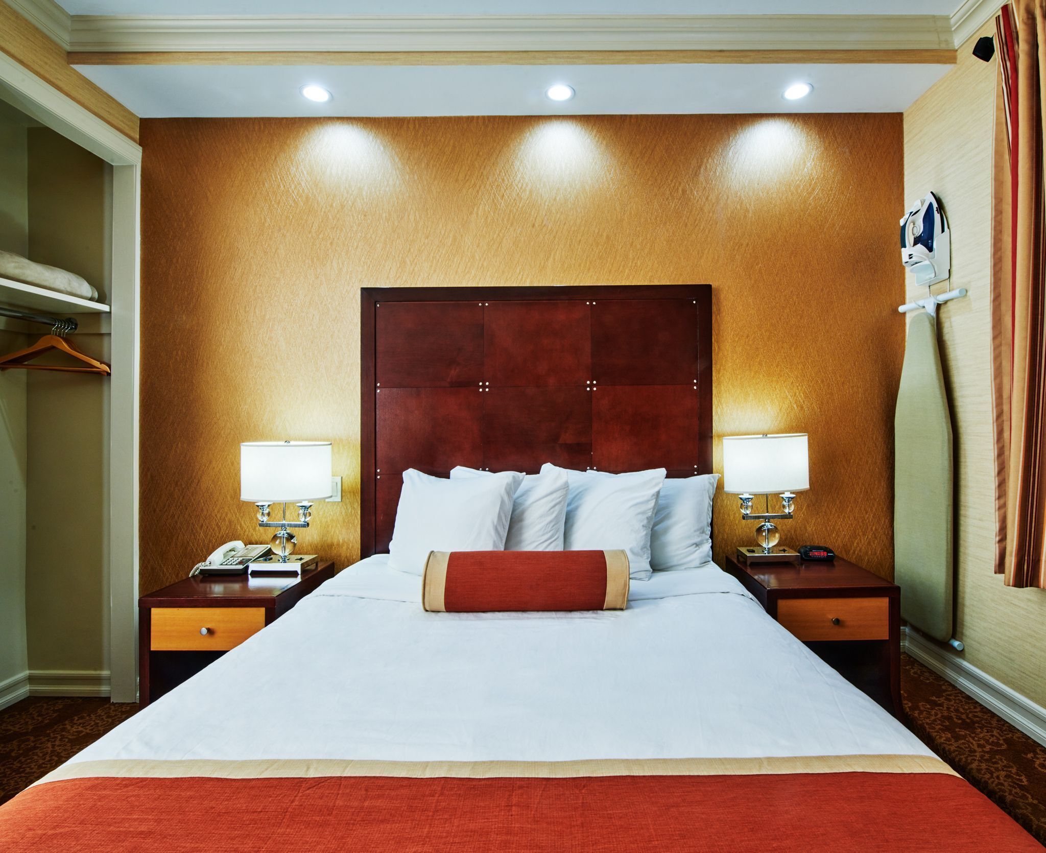 A hotel room with a large bed, two bedside tables with lamps, a wooden headboard, and a red throw pillow.