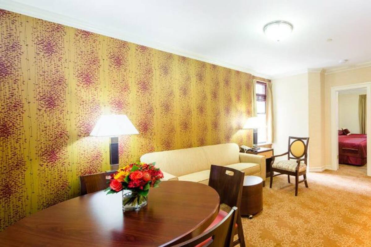 A hotel room with a dining area, a sofa, a chair, a table with flowers, a lamp, a bed, and a TV.