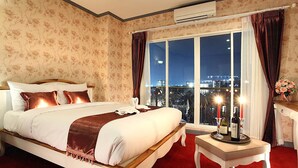 A hotel room with a large bed, a nightstand, a small table with a candle, and a view of the city at night. - Victoria Nimman Hotel (Chiang Mai)