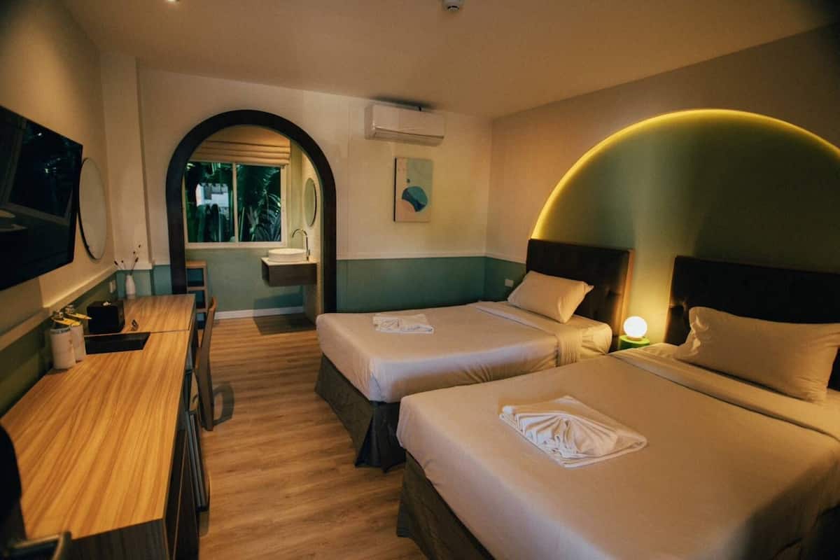 A hotel room with two beds, a desk, a mirror, and a bathroom with a sink and window.