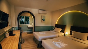 A hotel room with two beds, a desk, a mirror, and a bathroom with a sink and window.