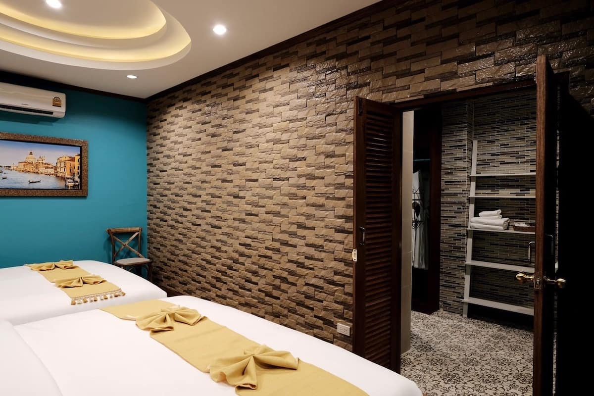 A hotel room with a brick wall, a bed with a beige bedspread, a wooden door, and a shelf with towels.