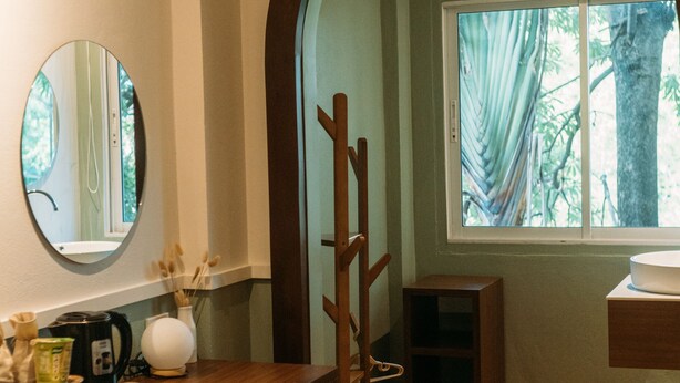A room with a wooden desk, a chair, a mirror, a window with curtains, and a view of trees outside. - Victoria Nimman Hotel (Chiang Mai)