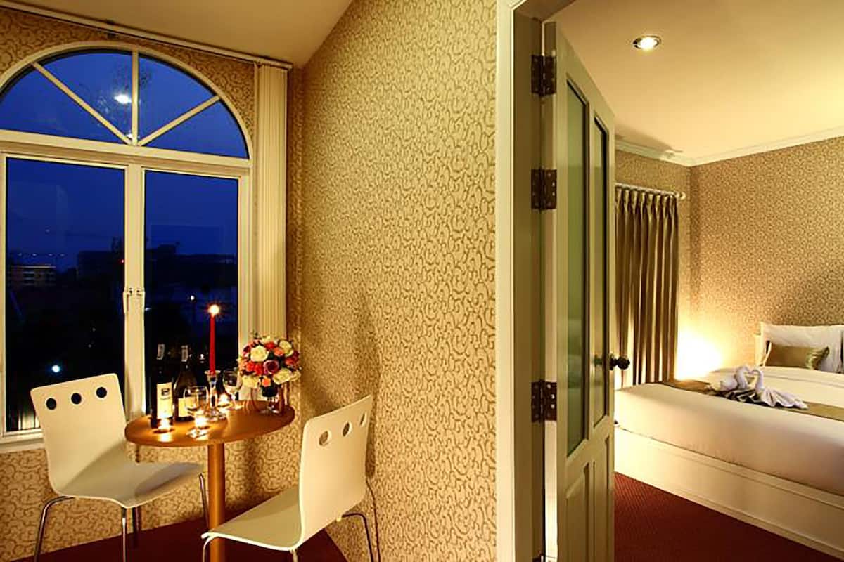 A hotel room with a large window, a dining area with a table and chairs, and a bed with a nightstand.