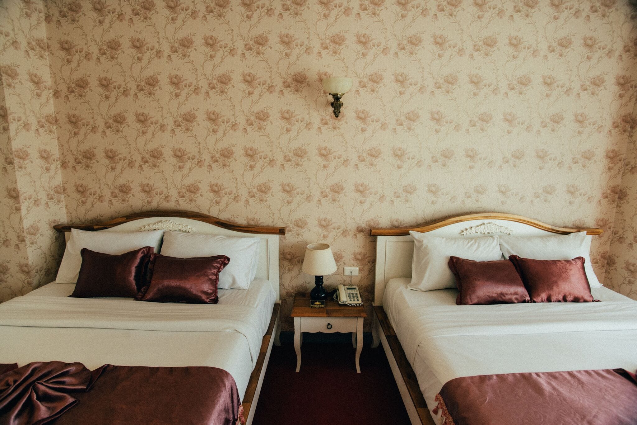 A hotel room with two beds, a nightstand with a lamp and phone, and floral wallpaper.