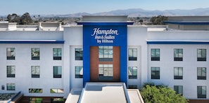 A hotel building with a prominent sign reading 'Hampton Inn & Suites by Hilton' and a view of distant mountains.
