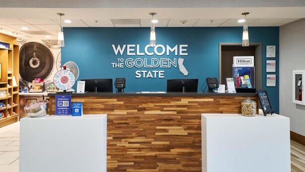A reception desk with a 'Welcome to the Golden State' sign, a fan, and a 'Hilton Honors' sign. - Hampton Inn & Suites Salinas (Salinas)