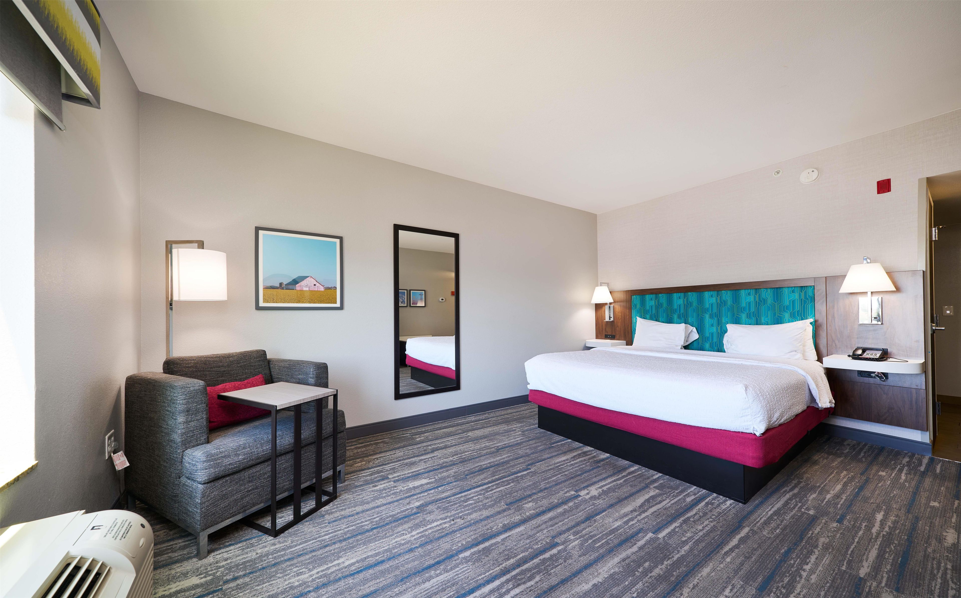 A hotel room with a bed, a chair, a desk, a mirror, and a picture on the wall.