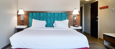 A hotel room with a large bed, two bedside lamps, a nightstand, a red accent pillow, and a wooden headboard.