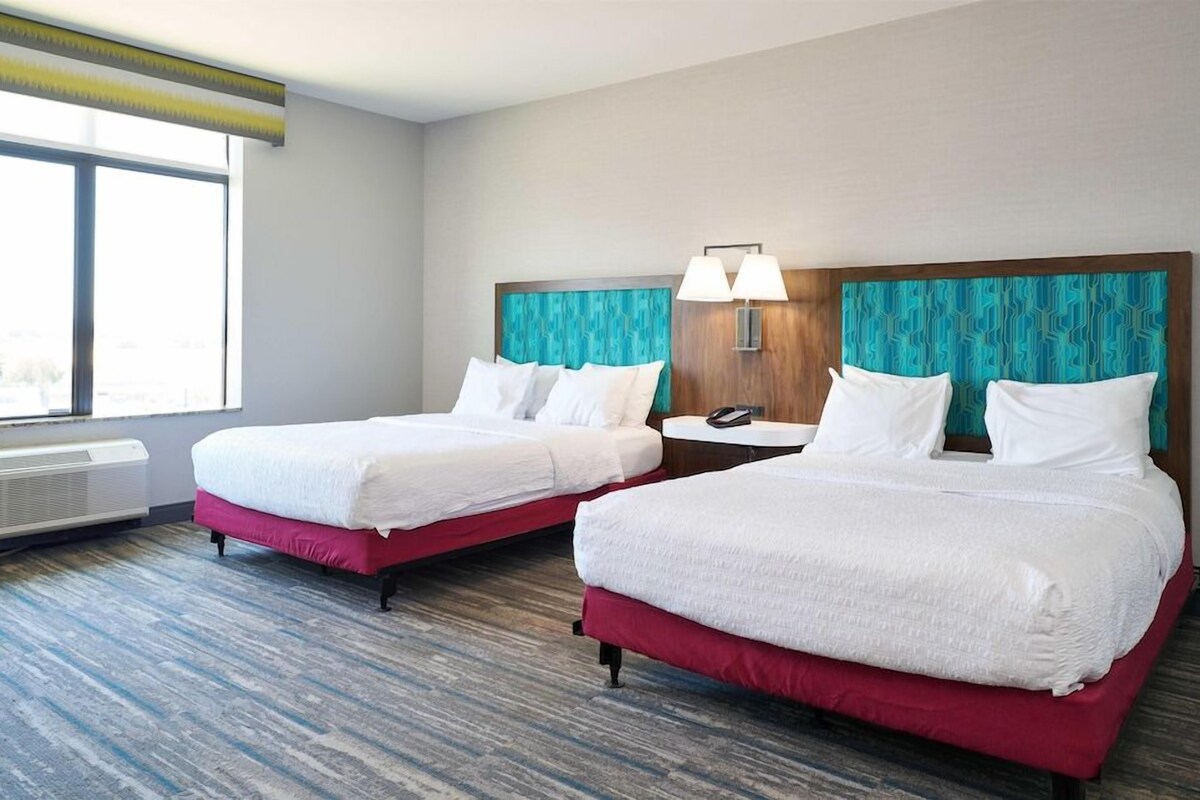 Habitación de hotel con dos camas, una ventana, un calefactor en la pared y una mesita de noche con lámpara.