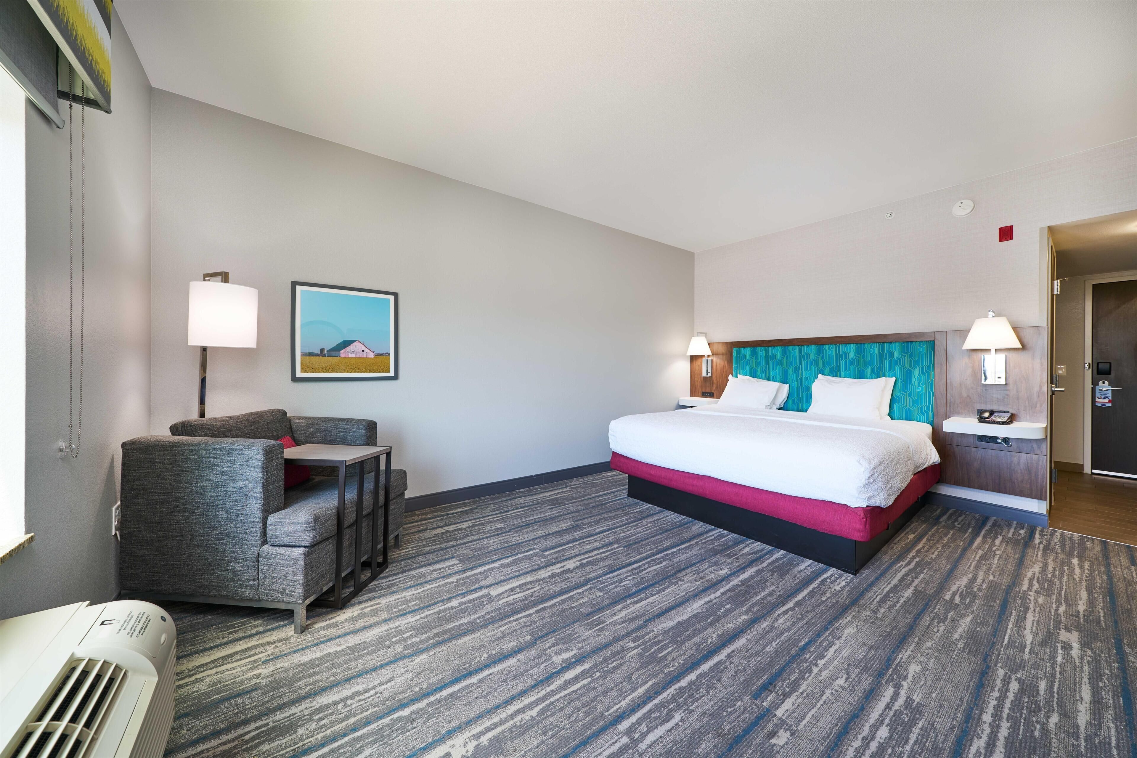 A hotel room with a bed, a desk, a chair, a picture on the wall, and a door.