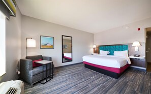 Habitación de hotel con cama, una silla, escritorio, espejo y un cuadro en la pared.
