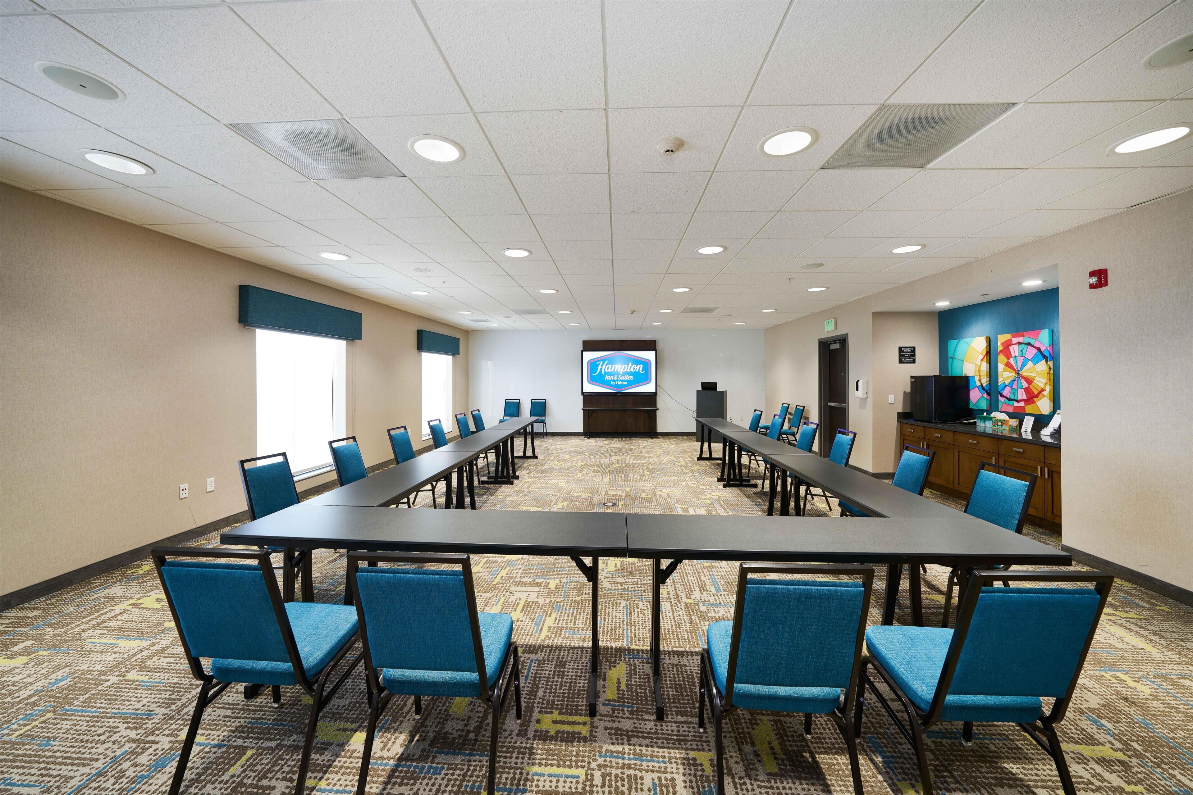 A conference room with a long table, blue chairs, a projector screen, and a colorful artwork on the wall.