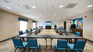 A conference room with a long table, blue chairs, a projector screen, and a colorful artwork on the wall.