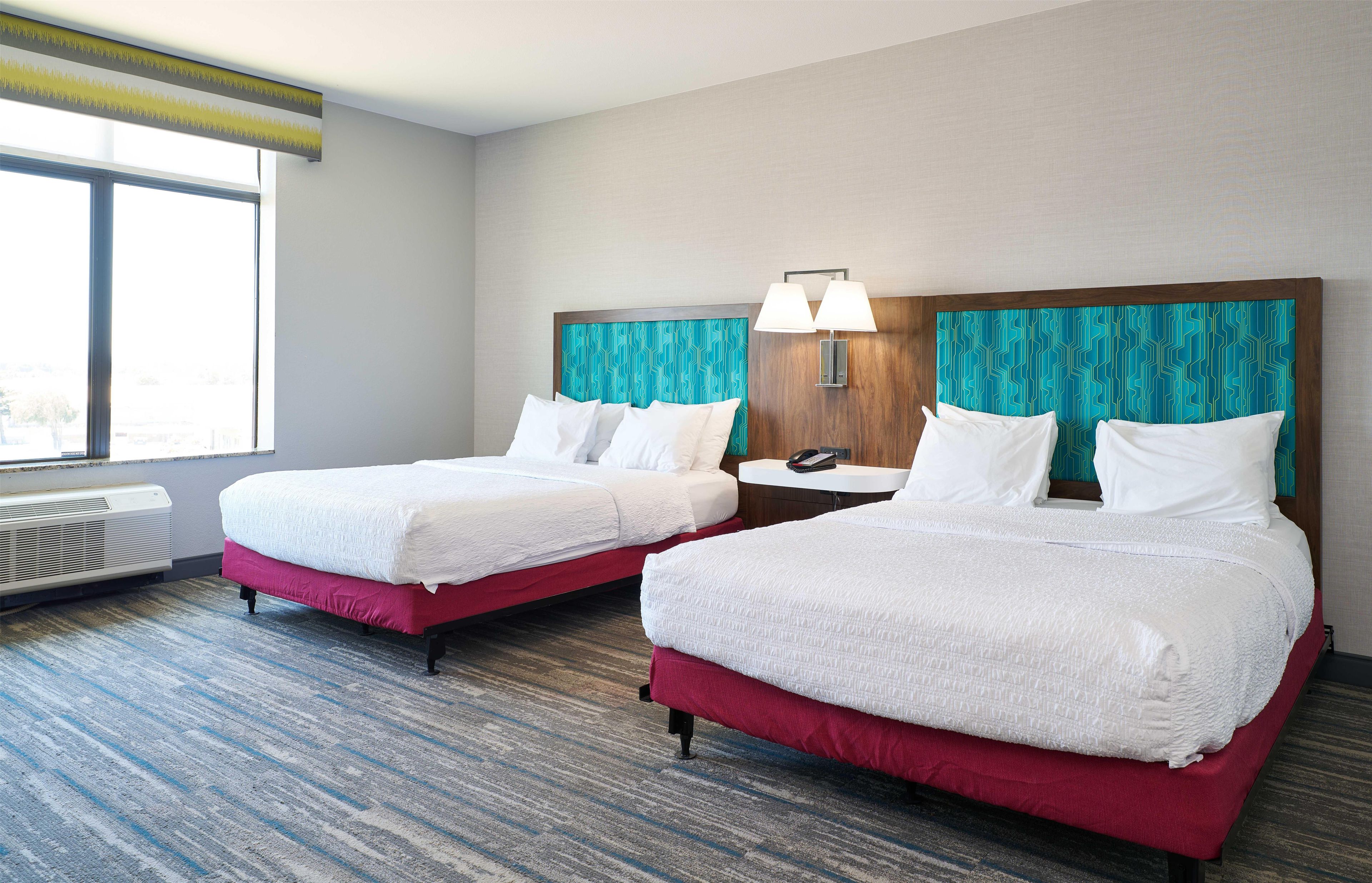 A hotel room with two beds, a nightstand, a lamp, and a window.