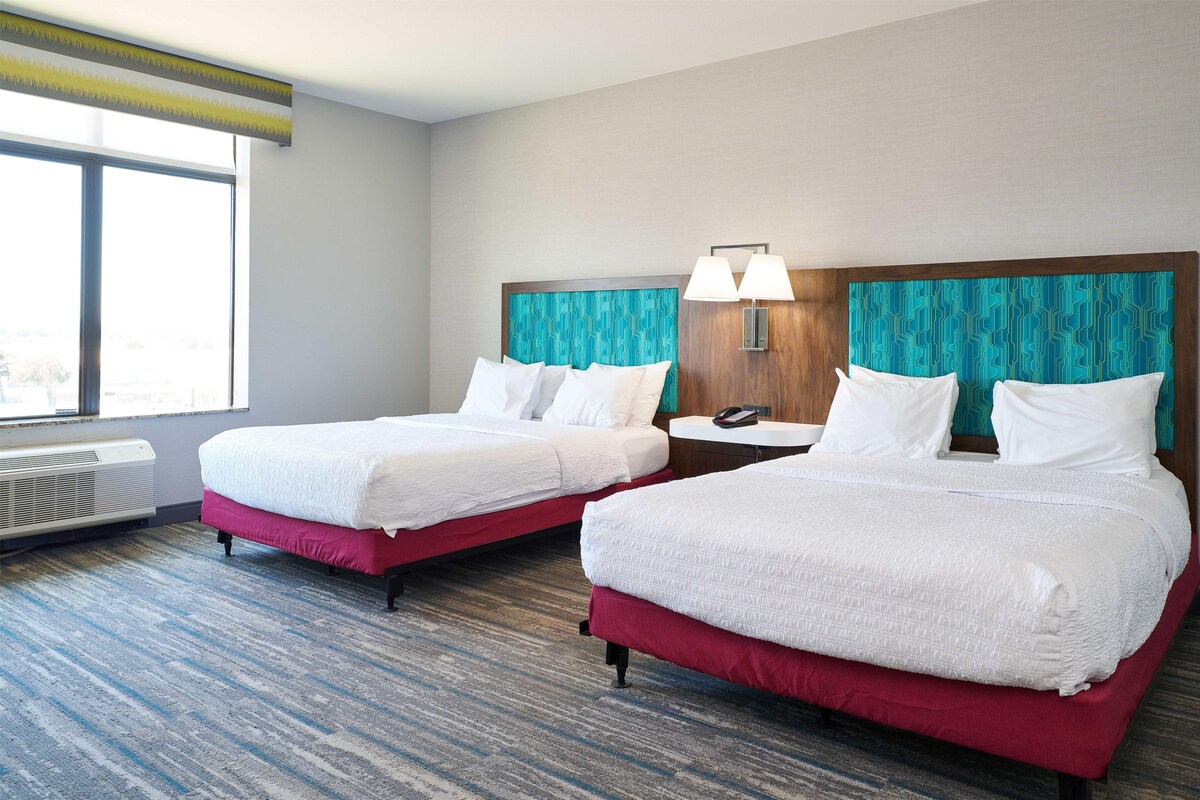 A hotel room with two beds, a nightstand, a lamp, and a window.