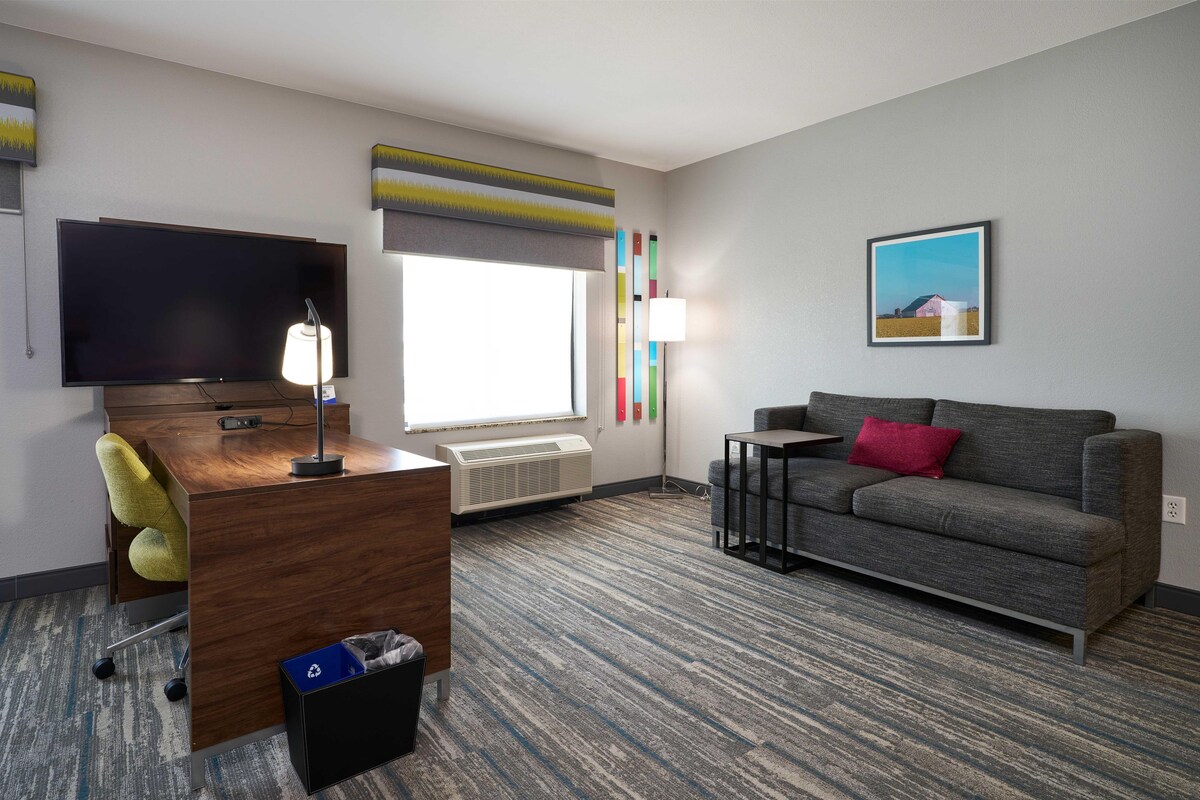 A hotel room with a flat-screen TV, a desk with a lamp, a grey sofa, a small table, a radiator, and a painting on the wall.