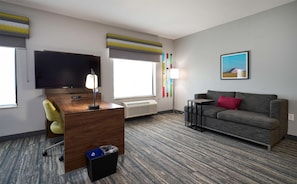A hotel room with a flat-screen TV, a desk with a lamp, a grey sofa, a small table, a radiator, and a painting on the wall.