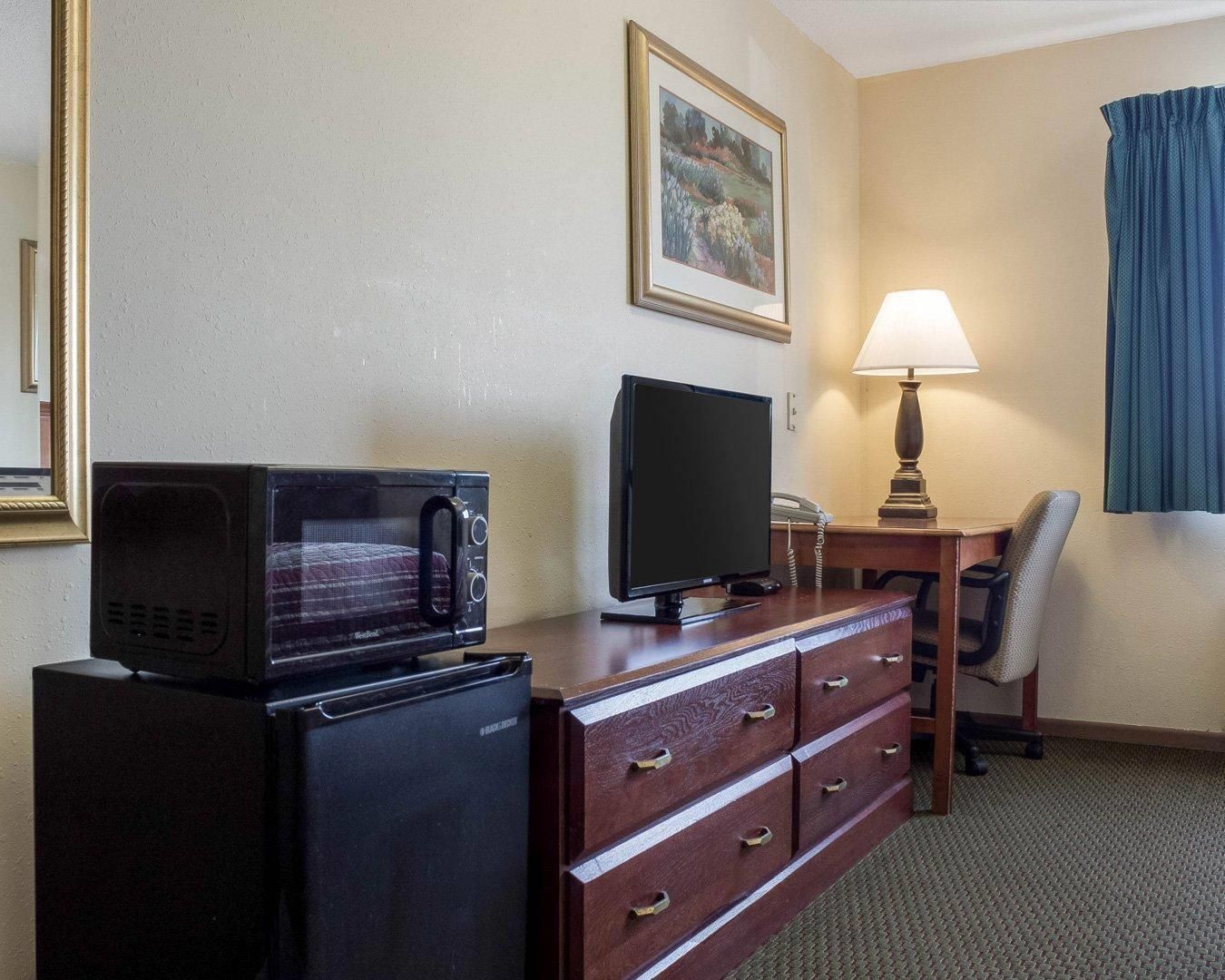 Habitación de hotel con microondas, televisor, escritorio con lámpara, silla y cómoda.