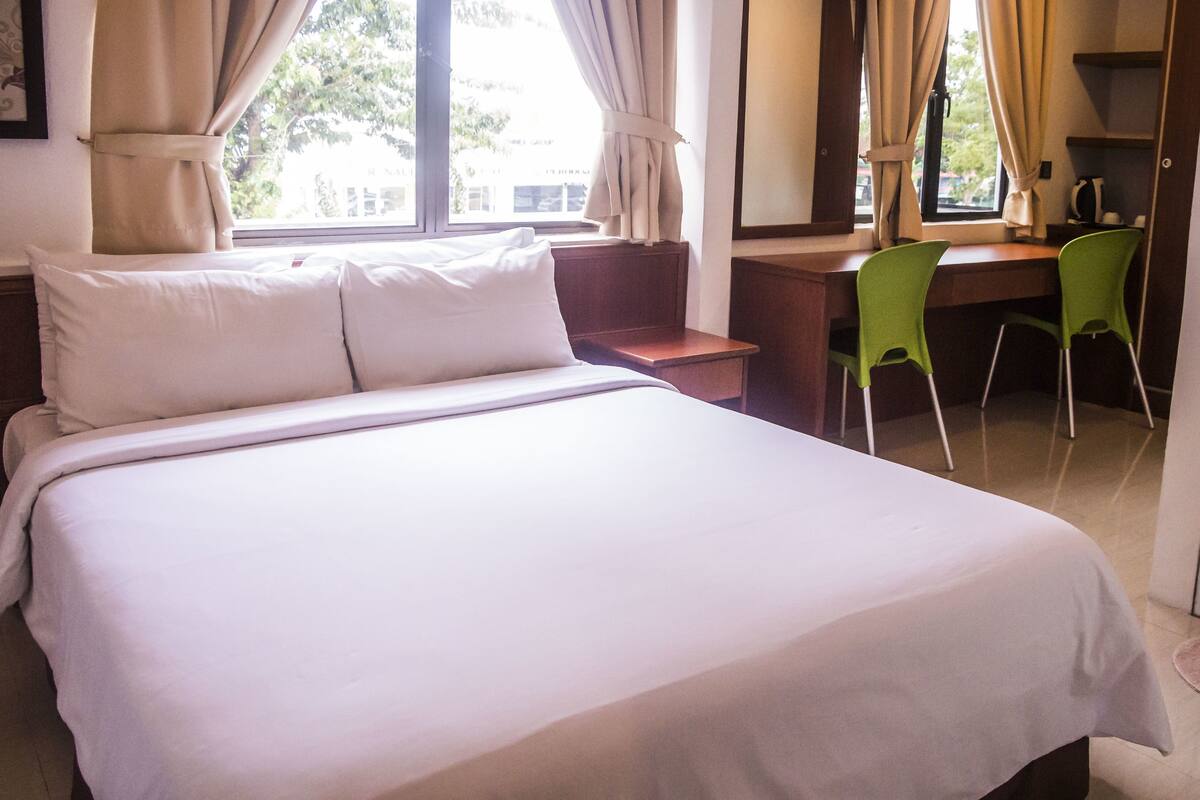A hotel room with a large bed, a desk, two green chairs, and a window with curtains.