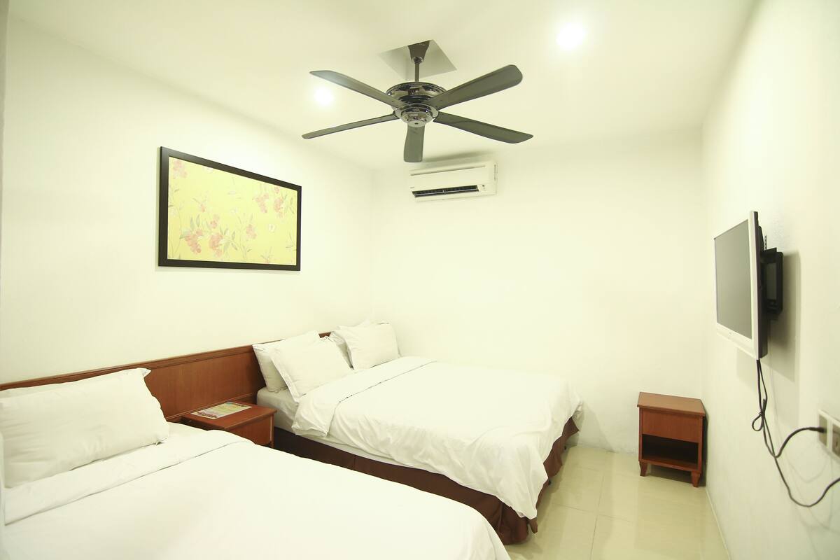 A hotel room with two beds, a ceiling fan, an air conditioning unit, a small bedside table, and a TV mounted on the wall.